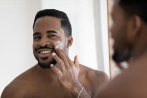 Young smiling African man reflect in mirror apply cleanse products on face or facial cream, put moisturizing or nourishing creme, enjoy soft hydrated skin. Skincare, personal hygiene routine concept