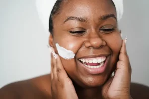 A black woman happily applying cream to her face, enhancing her skincare routine with a smile of contentment.