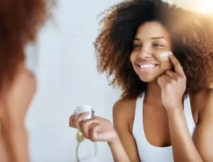 A black woman happily smears her face with skin care products, showcasing a range of beauty items on a table.