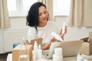 Smiling Internet shop employee holding skincare products and looking at laptop screen during video call