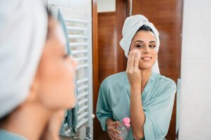 Beautiful young woman cleansing her face with a pad and removing her make up while looking in the mirror in the bathroom. november