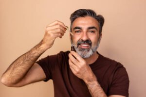 Smiling middle-aged man applying beard oil against a neutral beige backdrop